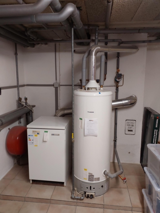 White vertical water heater in a utility room with a network of grey pipes overhead and a red expansion tank on the left.