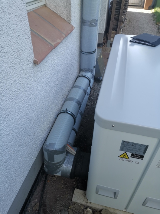 Outdoor air conditioning condenser unit installed in a narrow alley beside a textured wall, with insulated refrigerant pipes running along the wall