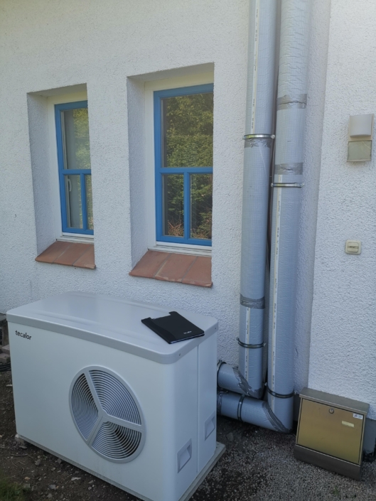 Outdoor air conditioning condenser unit with insulated pipes running up a white stucco wall beside two blue-framed windows.